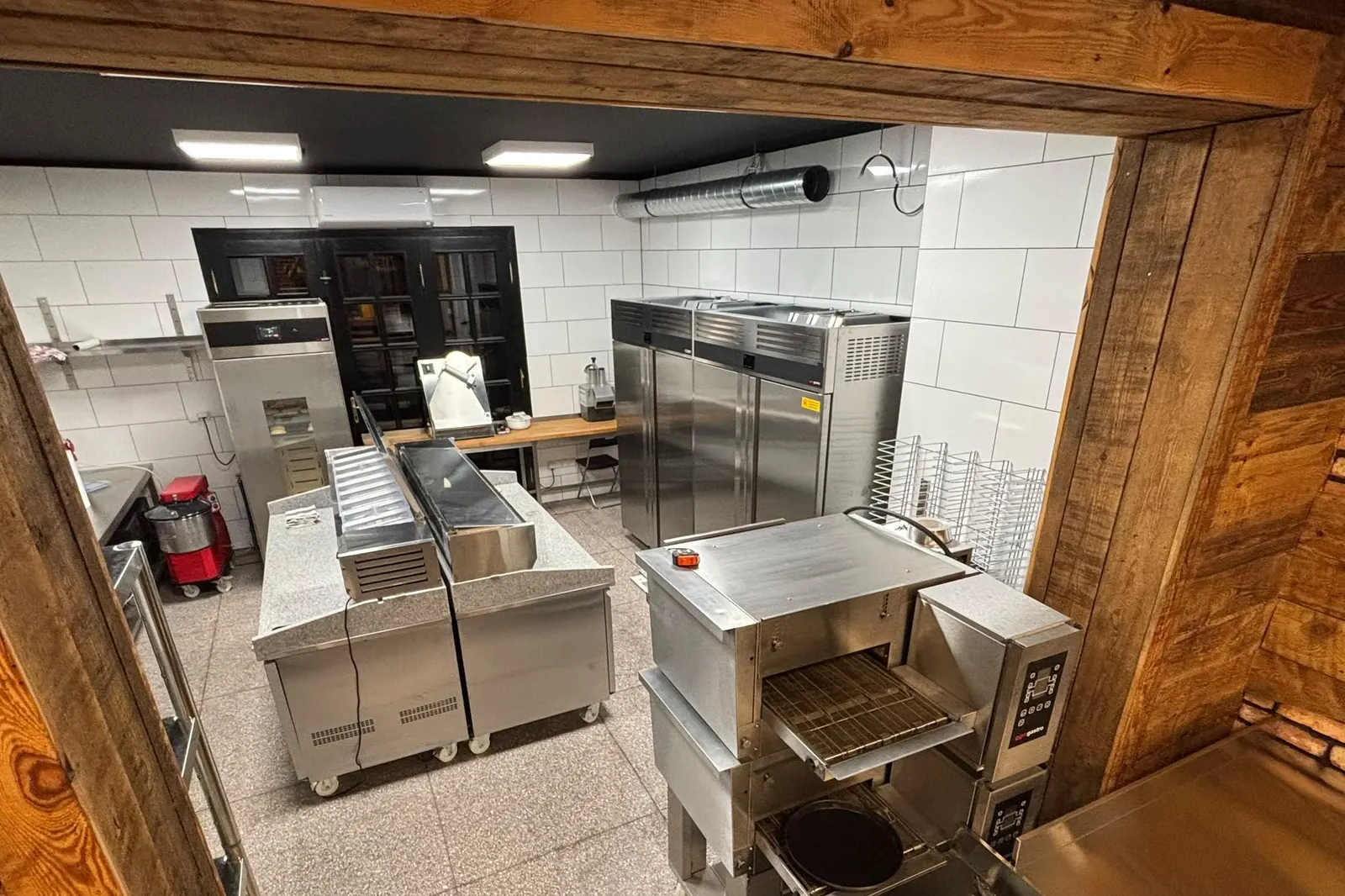 Kuchnia pizzerii z piecem tunelowym, widziana przez drewniane przejście